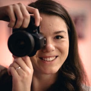 Junge Frau mit Dunkelbraunen Haaren, die in die Kamera lächelt und mit einer Hand eine Kamera hält.